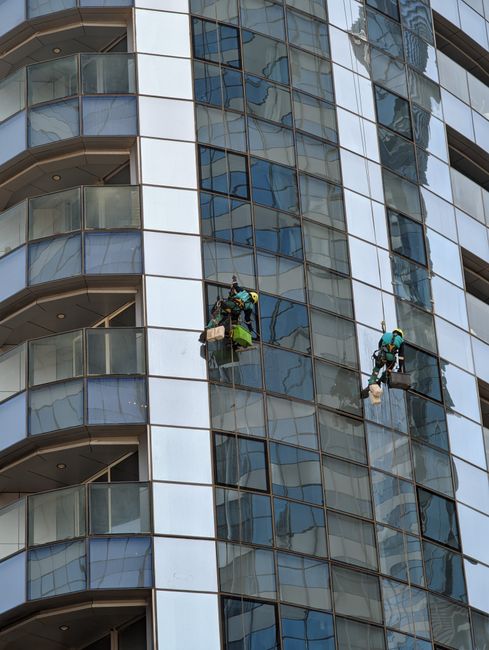 A professional window cleaner wearing a safety helmet and harness is cleaning the exterior glass facade of a modern high-rise building. The worker is seated on a platform suspended by ropes, using a s