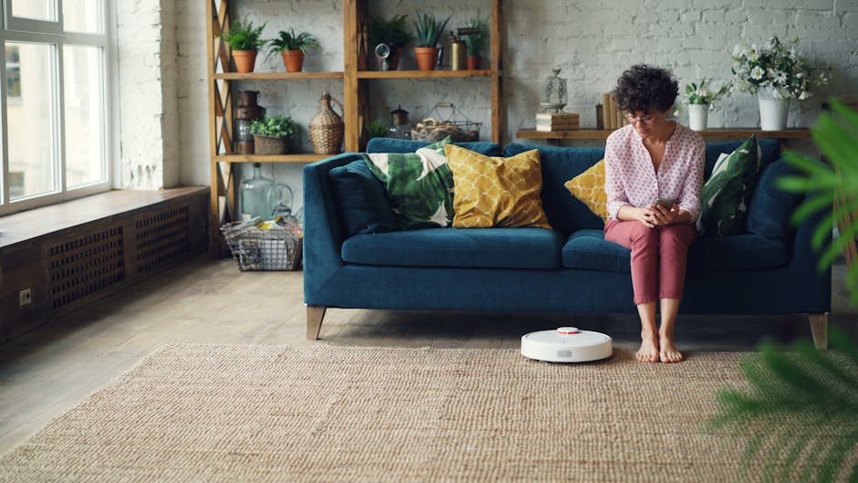 A woman with curly hair and glasses sits on a blue fabric sofa in a well-lit living room, looking at her mobile phone. The sofa is decorated with colorful cushions in yellow and green. In front of her