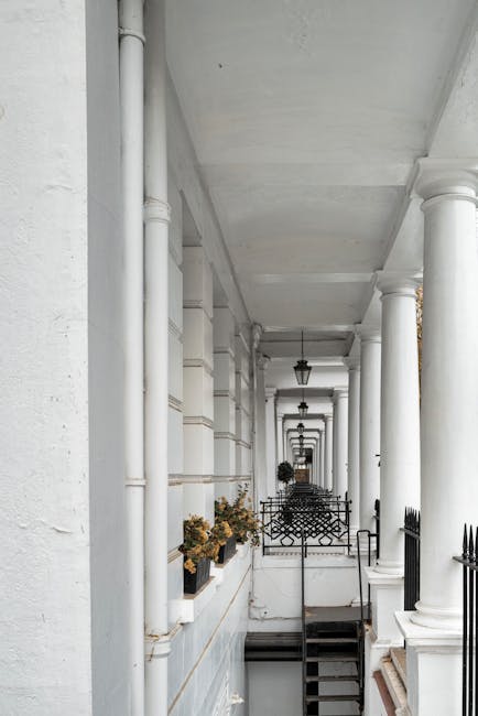 A long, narrow residential porch featuring white painted walls, multiple evenly spaced white columns supporting the ceiling, and black metal railings along the staircase. The floor appears clean with no visible dirt or debris, and the surface is likely stone or concrete. Small flower boxes with dried or faded plants are positioned along the wall, adding a touch of color to the predominantly white and black color scheme. Soft natural lighting illuminates the space, highlighting the smooth, well-maintained surfaces. This deep cleaning for Kensington townhouses near High Street Kensington emphasizes hygiene and aesthetic upkeep, consistent with professional domestic cleaning standards provided by Kensington Cleaner.