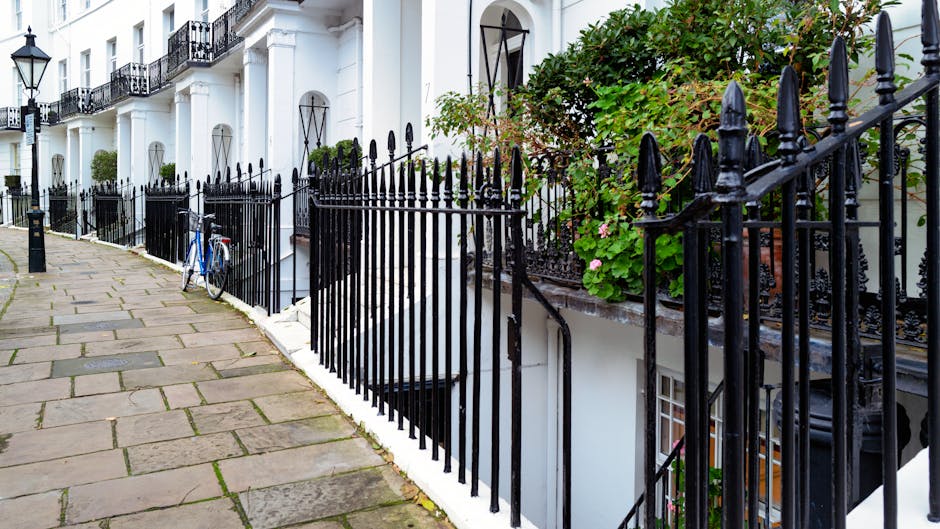 A row of white terraced townhouses near High Street Kensington, showcasing a paved sidewalk with large, rectangular stone slabs and a black wrought iron fence running along the front of the buildings. The fence features pointed finials and encloses small front gardens with green leafy plants and flowering shrubs. One townhouse has a slightly open door, and a blue bicycle is parked against the fence. The buildings have black iron balconies, white exterior walls, and large windows, with natural daylight illuminating the scene and highlighting the cleanliness and well-maintained appearance of the exterior surfaces. Kensington Cleaner specializes in deep cleaning and surface sanitisation for residential properties in this area.