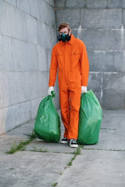 A sanitation worker in an orange safety uniform and safety helmet, viewed from above, is cleaning a sidewalk with a small broom and dustpan. The worker is collecting litter and debris, including paper
