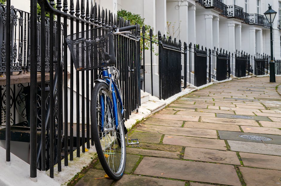 A residential sidewalk in a Kensington neighborhood, featuring large rectangular stone paving slabs with visible moss growth between some joints. To the left, a black wrought iron fence encloses a white building with classic architectural details, including white columns and black wrought iron balcony railings. A blue bicycle with a front basket is parked against the fence, partially leaning on it. The sidewalk is clean, with some fallen leaves and a few small cracks. Soft natural lighting highlights the gleaming surface of the paving stones and the ornate fence, illustrating a well-maintained outdoor area, as part of the interior of a property that Kensington Cleaner might service for deep cleaning and surface sanitation.